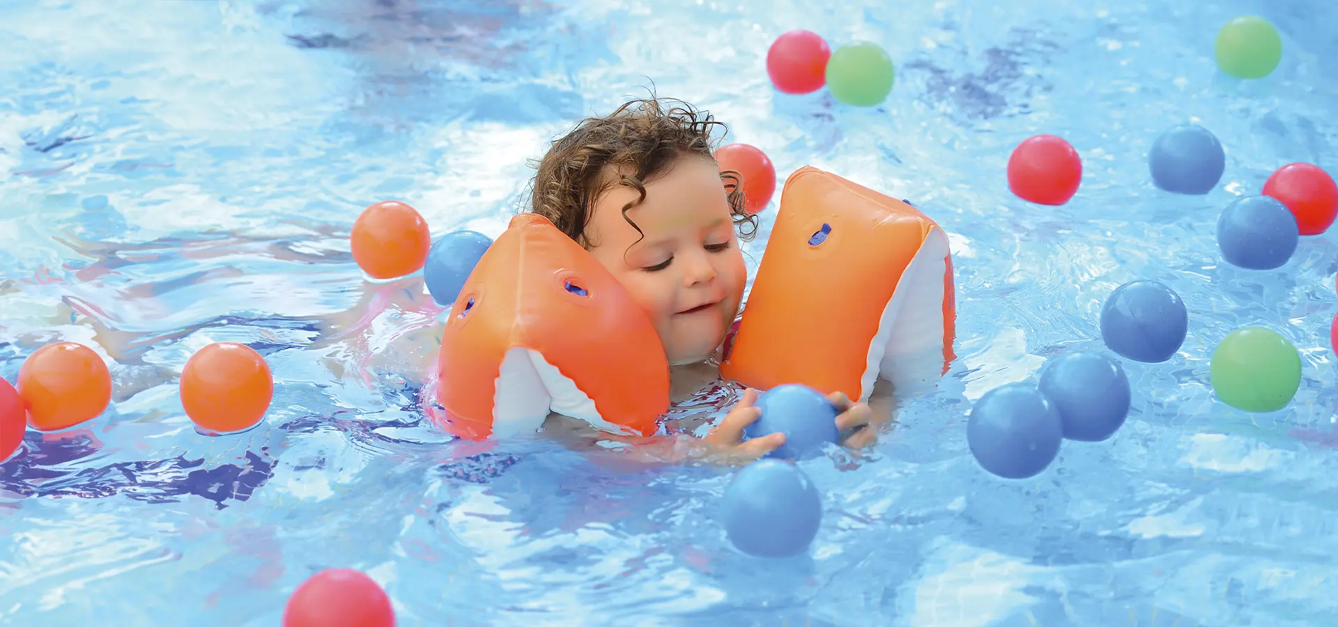 Kleines Kind mit Schwimmflügeln spielt im Wasser mit Bällen